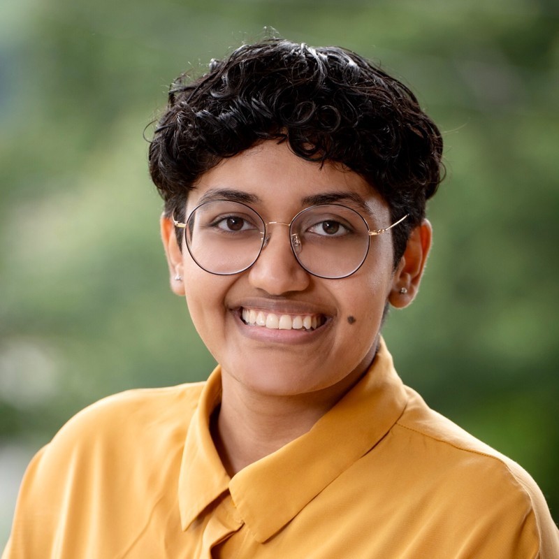 Chandu Avni smiles. They have dark, short, curly hair and brown skin. They are wearing glasses and a yellow, collared shirt.